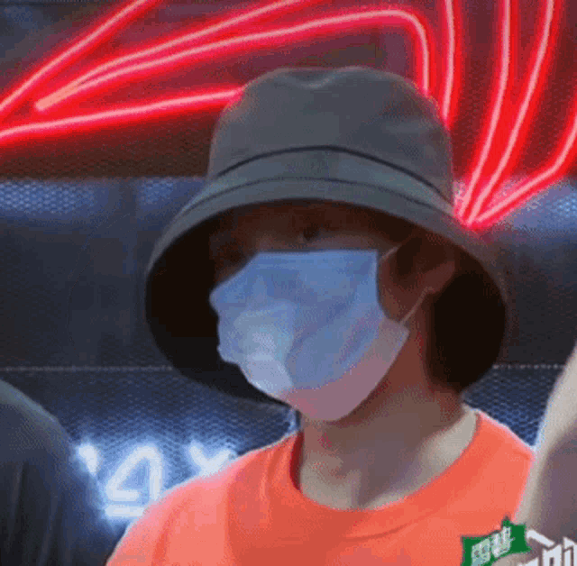 a man wearing a hat and a mask is standing in front of a red neon sign .