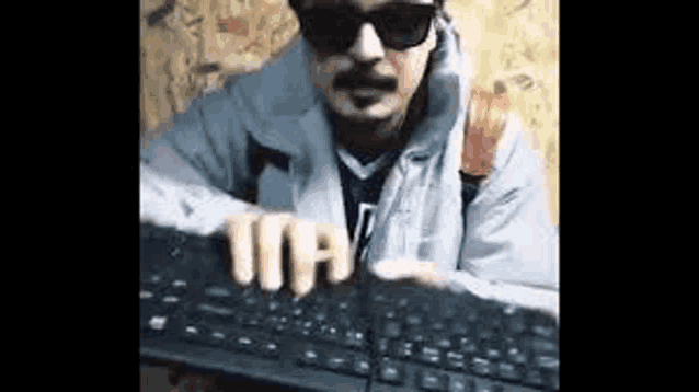 a man with a mustache and sunglasses is typing on a computer keyboard .