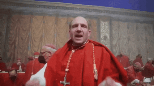 a man in a red robe with a cross around his neck is standing in front of a group of cardinals .
