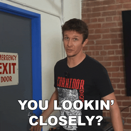 a man wearing a black shirt that says " you lookin ' closely " stands in front of an emergency exit door