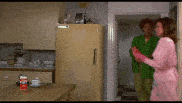 a woman in a pink robe stands in a kitchen next to a man in a green jacket