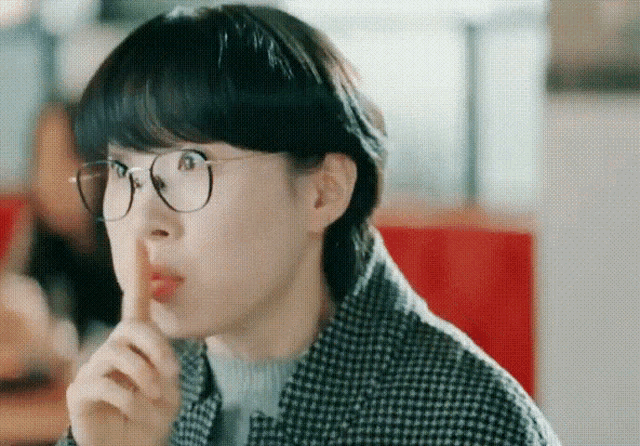 a woman wearing glasses and a plaid jacket is holding her finger to her mouth .