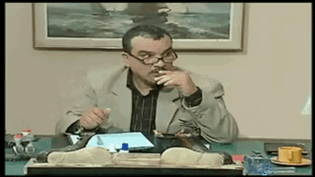 a man wearing glasses sits at a desk in front of a picture