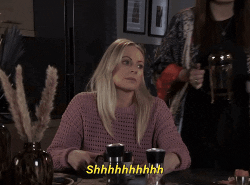 a woman in a pink sweater sits at a table with the word shhhh written in yellow letters