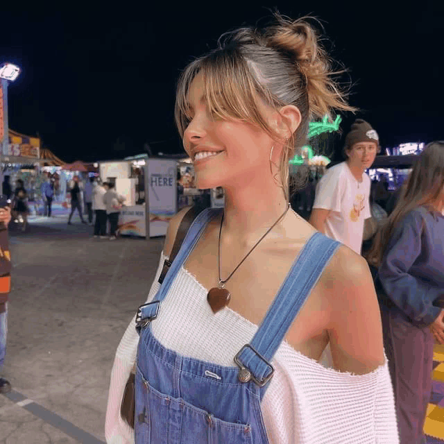 a woman wearing overalls and a white sweater smiles in front of a food stand that says here