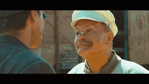a man with a mustache wearing a yellow hat talks to another man