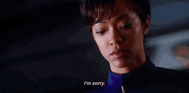 a woman in a blue shirt is saying i 'm sorry in a dark room .