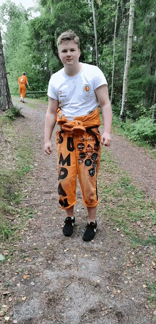 a young man wearing a white shirt and orange pants with the word su on them stands on a dirt path