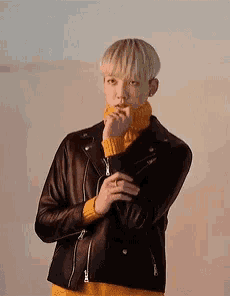 a young man wearing a black leather jacket and a yellow turtleneck is standing in front of a wall .