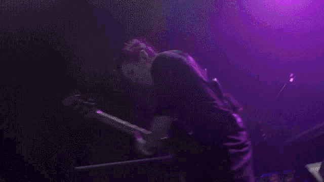 a man playing a guitar and singing into a microphone in front of purple lights