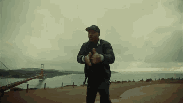 a man in a black jacket and black hat stands in front of a golden gate bridge