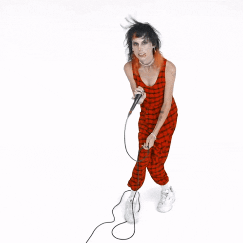 a woman singing into a microphone while wearing a red plaid jumpsuit