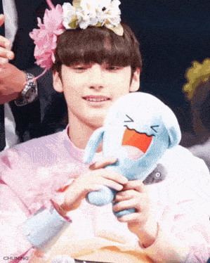 a young man with a flower crown on his head holds a stuffed animal