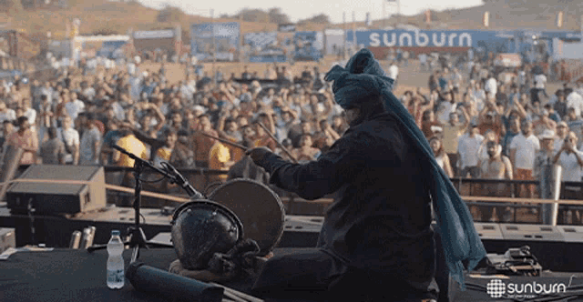 a man playing a drum in front of a crowd with a sunburn sign behind him