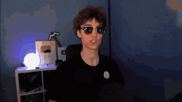 a young man wearing sunglasses and a black shirt is sitting in a chair in a dark room .