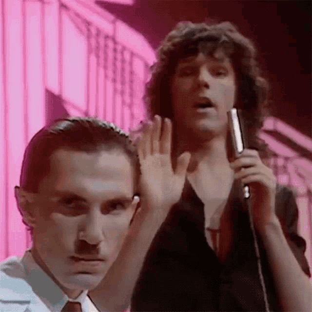 two men are standing next to each other and one of them is holding a microphone and waving .