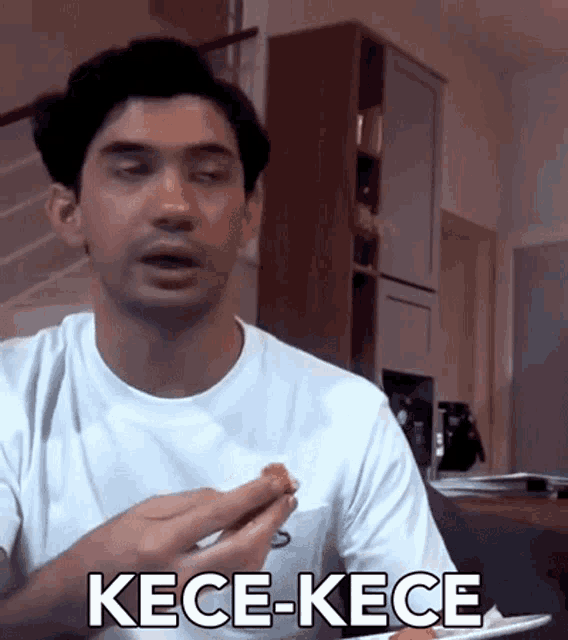 a man in a white shirt is holding a piece of food and the word kece-kece is on the bottom of his shirt