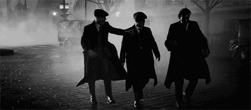 three men in suits and hats are walking down a cobblestone street at night .