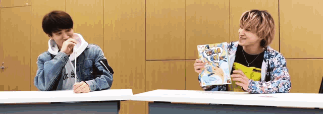 two young men are sitting at a table and one is holding a book that says ' superman ' on it