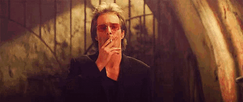a man in a suit and red sunglasses is smoking a cigarette in a dark room .