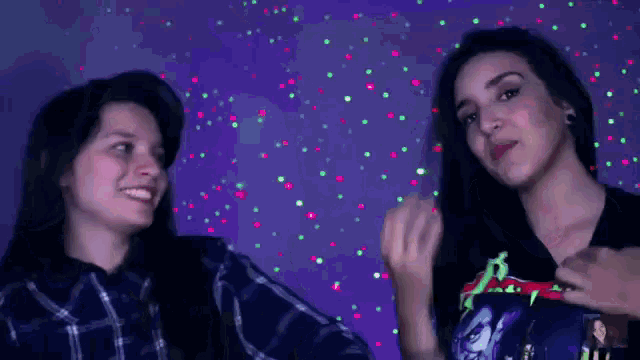two women are standing next to each other in front of a purple background with green and pink lights .