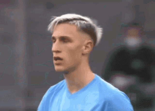 a soccer player with blonde hair is wearing a blue shirt and looking at the camera .