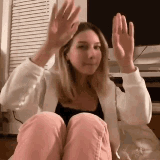 a woman is sitting on the floor with her hands in the air and waving at the camera .
