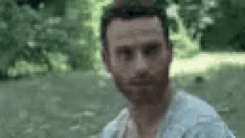 a man with a beard is standing in the grass in a park looking at the camera .