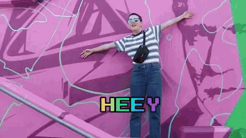 a woman is standing in front of a pink wall with her arms outstretched and the word heey written on the bottom .