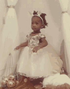 a little girl in a white dress with bunny ears standing next to a basket of easter eggs