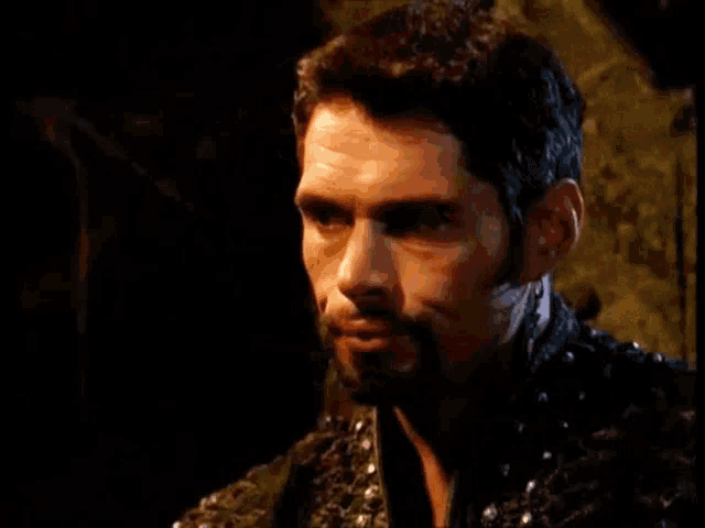 a man with a beard is sitting in a dark room talking to a woman named xena .