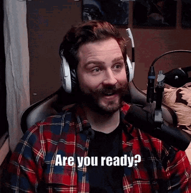 a man wearing headphones and a plaid shirt is sitting in front of a microphone and says are you ready ?