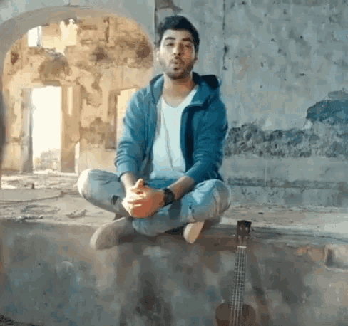 a man in a blue jacket is sitting on a wall with his legs crossed next to a guitar