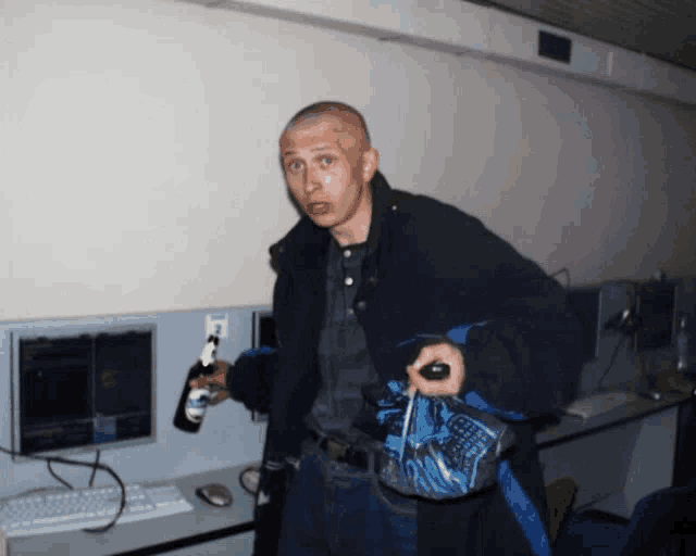 a man in a black jacket holds a bottle of beer in front of a computer