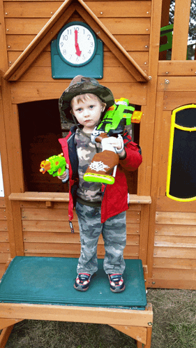 a little boy holding two toys in front of a wooden playhouse with a clock on the wall that says 1:11