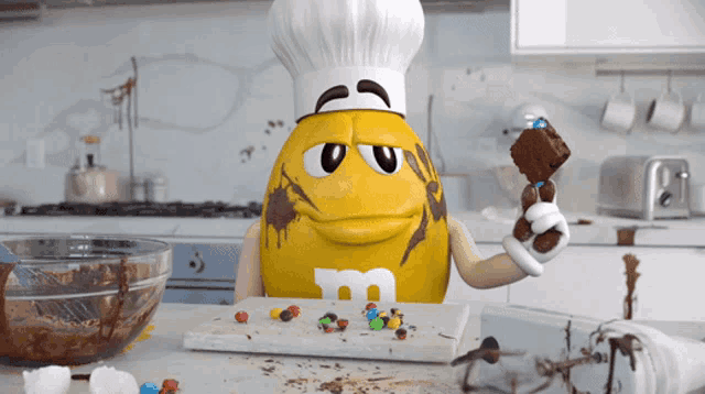 a yellow m & m holding a brownie and wearing a chef hat