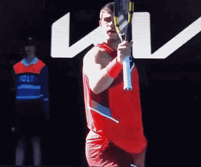 a man in a red shirt is holding a tennis racket in front of a kia logo