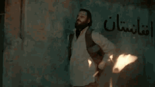 a woman is holding a gun in front of a wall with arabic writing .