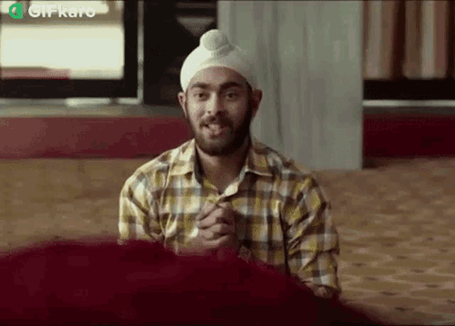 a man wearing a turban and a plaid shirt is sitting in a room with his hands folded .