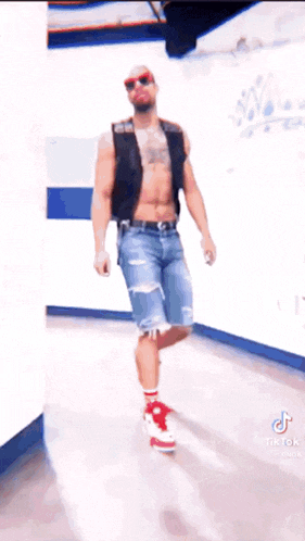 a man without a shirt is standing in a hallway wearing red shoes and sunglasses .