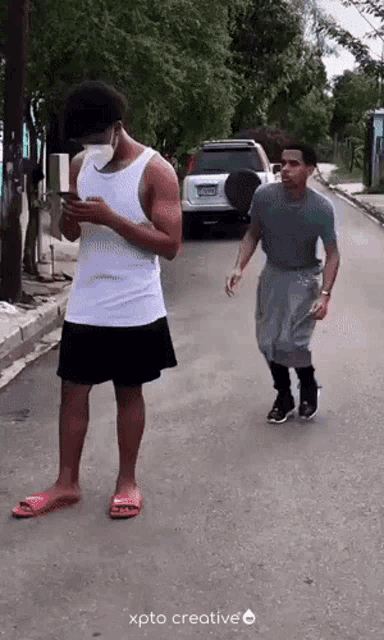 a man wearing a mask looks at his phone while another man walks down a street