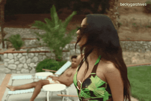 a woman in a green swimsuit stands in front of a man laying on a lawn chair