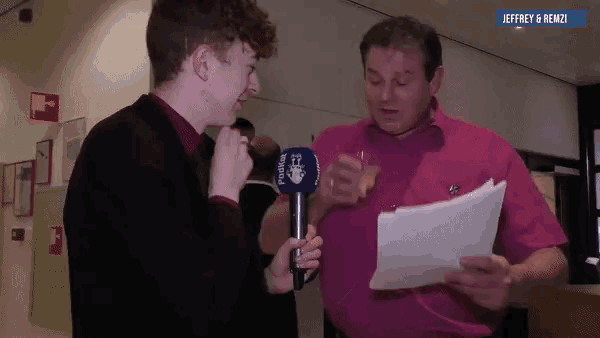 a man in a pink shirt is holding a piece of paper and talking to another man in a black suit