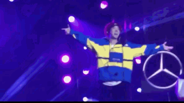 a man in a yellow and blue jacket is standing on a stage with purple lights behind him