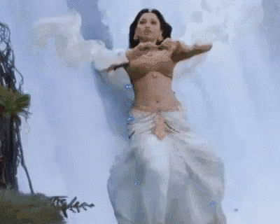 a woman in a white dress is standing in front of a waterfall with her arms outstretched .