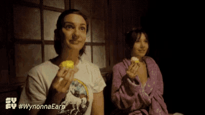 two women are sitting next to each other and eating cupcakes in a dark room .