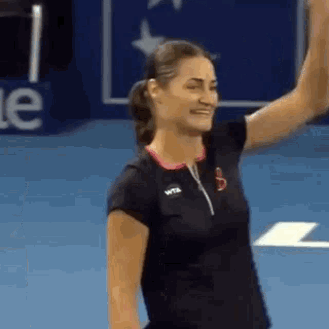 a woman in a black shirt is standing on a tennis court raising her hand .
