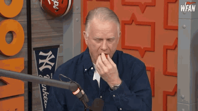 a man in a blue shirt is eating something in front of a microphone with a ny yankees banner behind him
