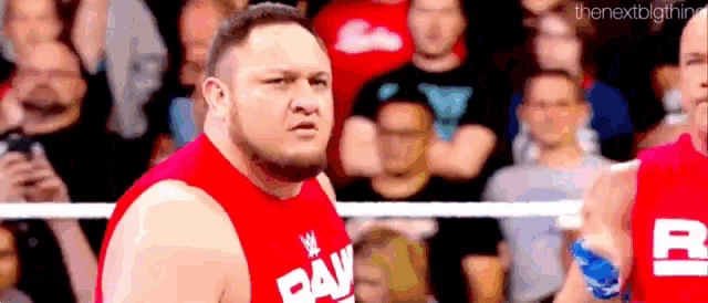 a wrestler in a red shirt is standing in a wrestling ring in front of a crowd .