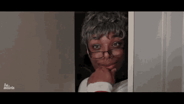 a woman wearing glasses and a wig is peeking out of a doorway and smiling .